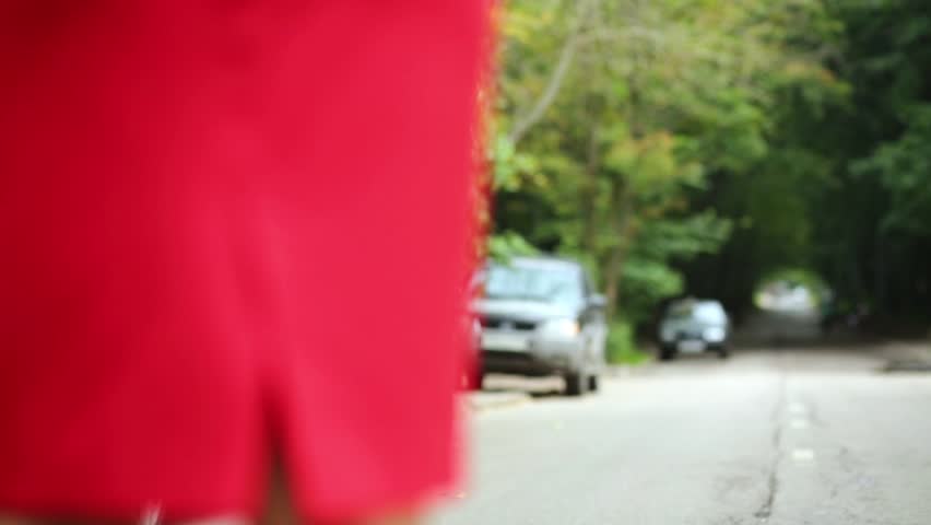 Young girl dressed in red skirt and blue blouse walks away along road with parked cars.