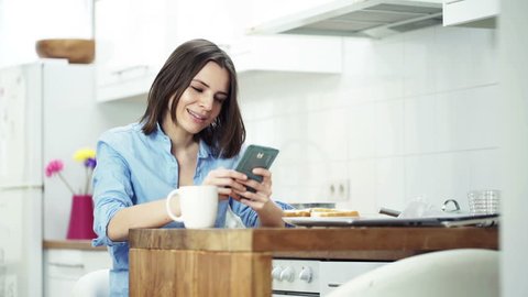 Happy, young woman using smartphone and drinking coffee by table in kitchen 
