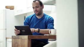 Man chatting on tablet computer and drinking coffee by table in kitchen
