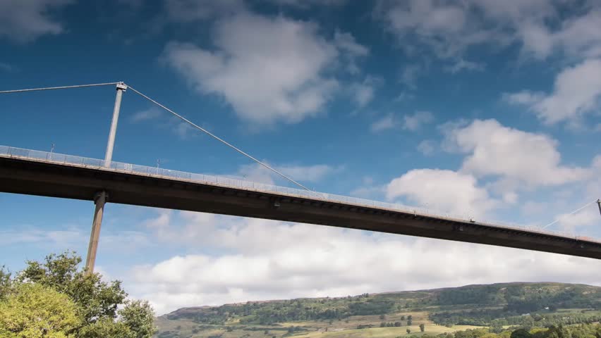 Time-lapse of Erskine Bridge in Renfrewshire near Glasgow, Scotland. Pan from left to right as clouds rush across a blue sky. Road bridge spans the River Clyde. Hills are in the background.