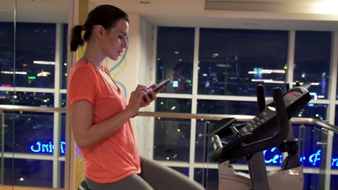 Young woman with smartphone riding stationary bike in gym at night
