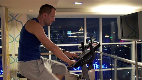 Young man with smartwatch riding stationary bike in gym at night

