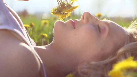 Beauty Girl lying on dandelions meadow and smelling flowers. Beautiful Spring Young Woman Outdoors Enjoying Nature. Healthy Girl in Green Grass. Allergy free. Slow motion 240 fps. 1080p