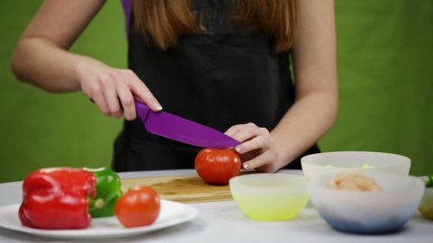 Person cut tomato on cutting board. Female person with ceramic knife slicing red tomato on cutting board with vegetables in front. Green background.