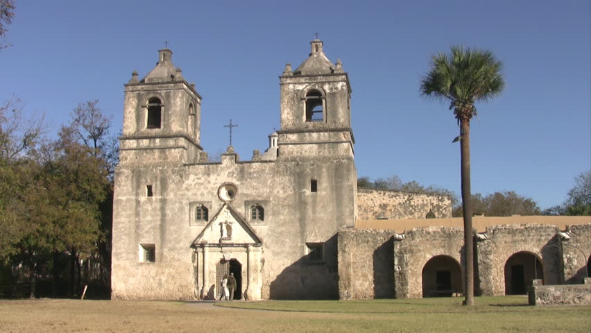 Video of old Spanish Mission Concepcion San Antonio, Texas. Early 1700s by missionaries from Spain. Bell towers, cathedral, rock well and stone walls. People entering through carved wooden door.