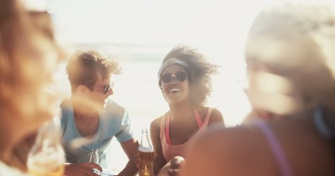 Laughing mixed racial group of friends drinking beer and relaxing together on the beach in slow motion