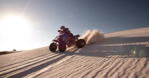 Competitive quad bike racer kicking up sand while driving up a sand dune on a summer evening with sun flare, Slow Motion, Panning