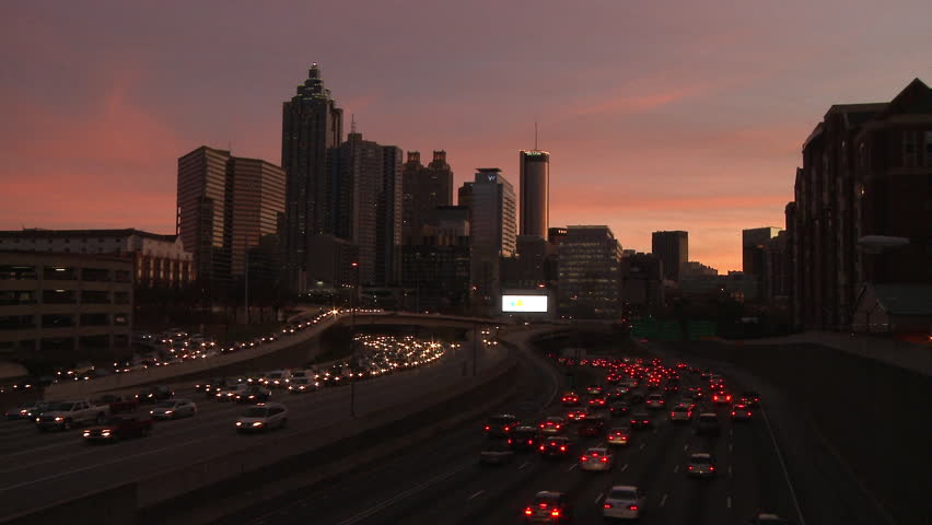 Static, wide, nighttime shot of the Atlanta Skyline with traffic below. Red brake lights and headlights can be seen. Traffic is going both ways. Lights from cars illuminate the scene.