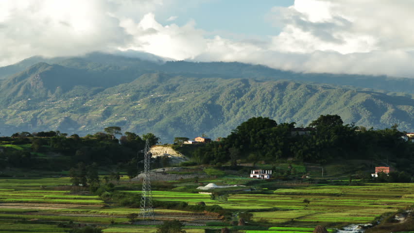 Time-lapse of the countryside of Nepal with changing light and billowing clouds over mountains. The valley floor is covered in fields with a few buildings. Cropped.