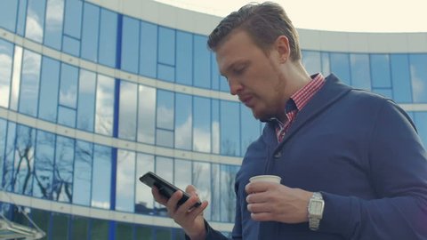 Attractive businessman with coffee in hand uses a smart phone. He stands in front of an office building and looks very serious and concerned