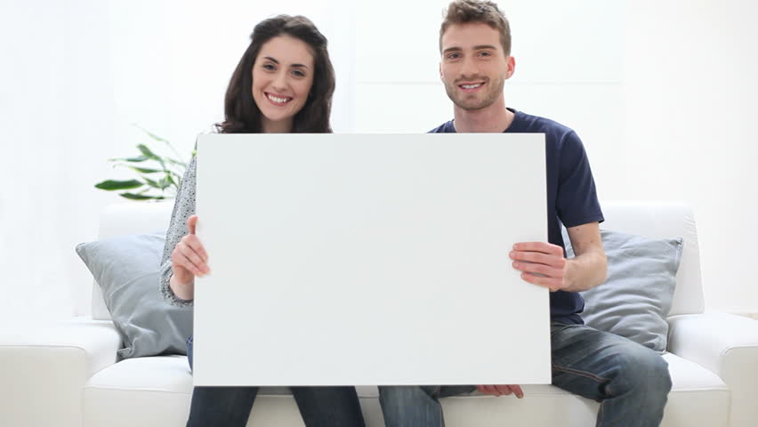 Portrait Of Smiling Young Couple Holding Placard At Home. Happy girl and smiling guy looking at camera with blank sign. Happy couple sitting on couch an holding a white placard. Royalty-Free Stock Footage #9798860