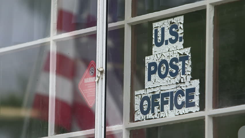 A US flag is reflected in the window of this small rural post office, with faded lettering.