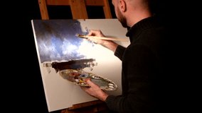 Stylish, bald, bearded painter in dark sweater goes on drawing a new painting with oil paints holding the palette in his hand on easel, black background, back light