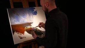 Bald, stylish, bearded painter in dark sweater goes on drawing a new painting with sky and ground by oil paints on easel holding the palette in his hand on black background, back light, slow motion