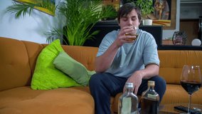 Home alone drowning in alcohol. Slow motion total RAW footage of a man drinking alone at home and seating on the couch and is bored.