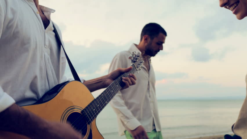 Group of four friend enjoy themselves playing guitar and dancing together outdoor at beach sea-side in summer day at sunset - gimbal steadicam HD video footage