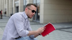 Handsome man  in blue shirt and sunglasses, sitting near building and reading notes. He is talking to someone.