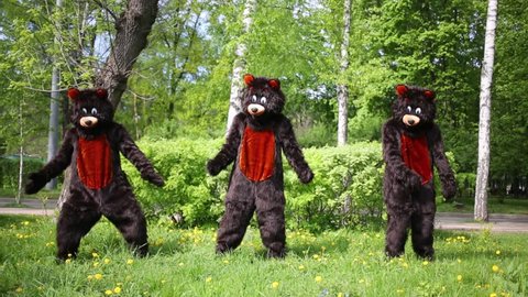 Three actors dressed in bear suits dance on grassy lawn at park.