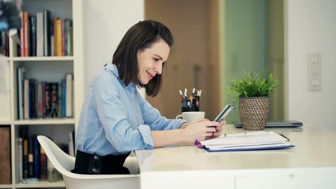 Young, pretty businesswoman with smartphone sitting by table at home
