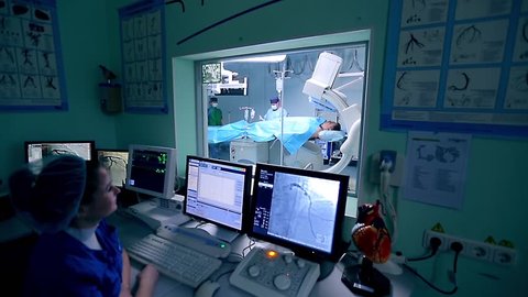 Assistant controls a cardio surgery in front of monitors in a control room. In an operation room a surgeon makes manipulations with a patient.