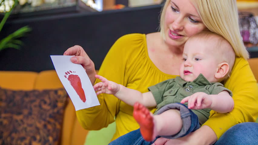 Mother showing the feet print to a small boy. Mother with a son seating on her lags is showing just made feet print to the son in living room, footage in slow motion.