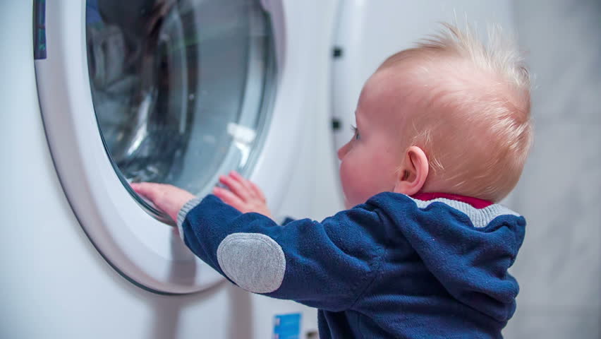 Clothes are washing in a washer. Young baby boy is seating next to the washer and looking haw the clothes are washing out. Royalty-Free Stock Footage #9999260